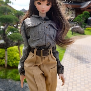 Puede incluir: Una muñeca con el pelo largo y castaño que lleva una camisa gris abotonada y pantalones cargo marrones con un cinturón marrón.