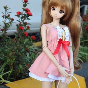 May include: A doll with long brown hair in pigtails wearing a pink dress with a white ruffle and a red bow. The doll is holding a wicker basket.