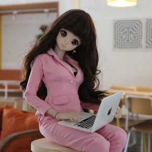 Op de afbeelding: Een pop met een roze blazer en broek zit op een houten kruk en gebruikt een zilveren laptop.