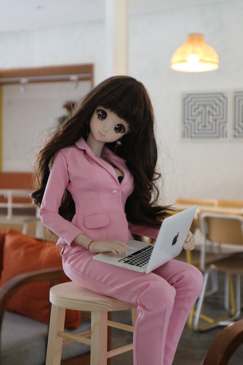 Op de afbeelding: Een pop met een roze blazer en broek zit op een houten kruk en gebruikt een zilveren laptop.