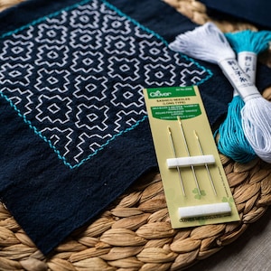 May include: A blue fabric square with a white geometric pattern stitched on it. The fabric is surrounded by a woven wicker mat. There are two spools of embroidery floss, one white and one teal, and a pack of three gold embroidery needles labelled "Clover Sashiko Needles (Long Type)".