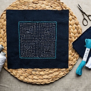 Peut inclure: Un carré de tissu bleu avec un motif de broderie blanc et turquoise. Le tissu est sur un tapis de paille tressé. Il y a deux bobines de fil, une blanche et une turquoise, et un paquet d'aiguilles.