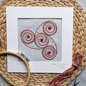 Op de afbeelding: Een ingelijst borduurwerk met een witte achtergrond en een beige linnen stof met een rood en bruin geborduurd ontwerp van drie in elkaar grijpende spiralen. Het stuk is gesigneerd met "Celtic Triskele, April '21".