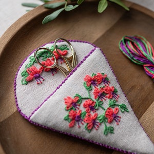 May include: A white linen scissor keeper with a purple trim and embroidered pink flowers. The keeper is filled with gold scissors.