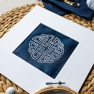 May include: A white framed embroidery hoop with a blue fabric background featuring a white Celtic knot design.