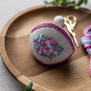 May include: A small, gray linen pincushion with a pink trim and a floral embroidery design in shades of pink, blue, and green. The pincushion is sitting on a wooden tray.