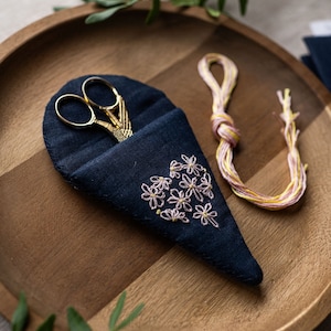 May include: A navy blue scissor case with a floral embroidery design, holding gold scissors. A skein of yellow and pink thread sits next to the case on a wooden tray. Craft supplies.