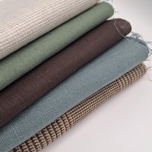 May include: A stack of five folded fabric swatches featuring various textures and colours. The top piece is a cream textured weave, followed by sage green, dark brown, dusty blue, and a bottom piece with a brown and cream checkered pattern.