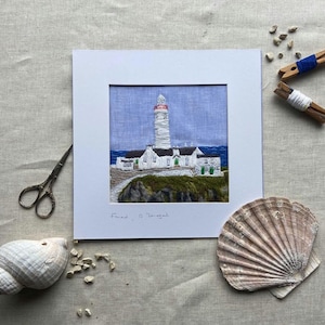 May include: A white framed embroidery of a lighthouse on a blue background. The embroidery is titled "Fanad, Co Donegal".