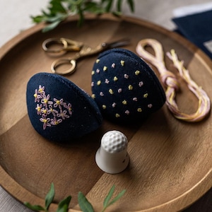 May include: Two blue fabric pincushions with embroidered floral designs, a white ceramic thimble, and a spool of pink and yellow thread on a wooden tray.