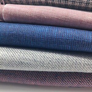 May include: A selection of fabric swatches in a range of colours, including grey, blue, pink, and black. The fabrics display diverse textures and patterns, such as herringbone and woven styles. The swatches are stacked, highlighting the variety of textiles.