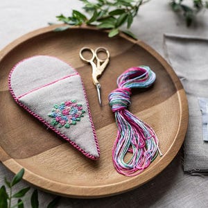 Puede incluir: Un estuche para agujas con forma de corazón con costuras rosas y un diseño de bordado floral. El estuche está en una bandeja de madera con un par de tijeras doradas, un carrete de hilo de bordar rosa y azul y un trozo de tela de lino.