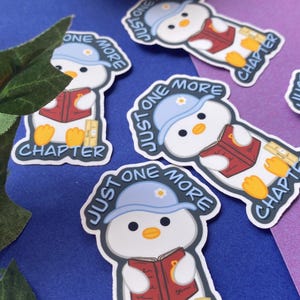 May include: A collection of stickers featuring a cartoon penguin reading a book. The penguin wears a blue hat with the text "Just One More" and the word "Chapter" is also visible. The stickers are on a blue and purple background.
