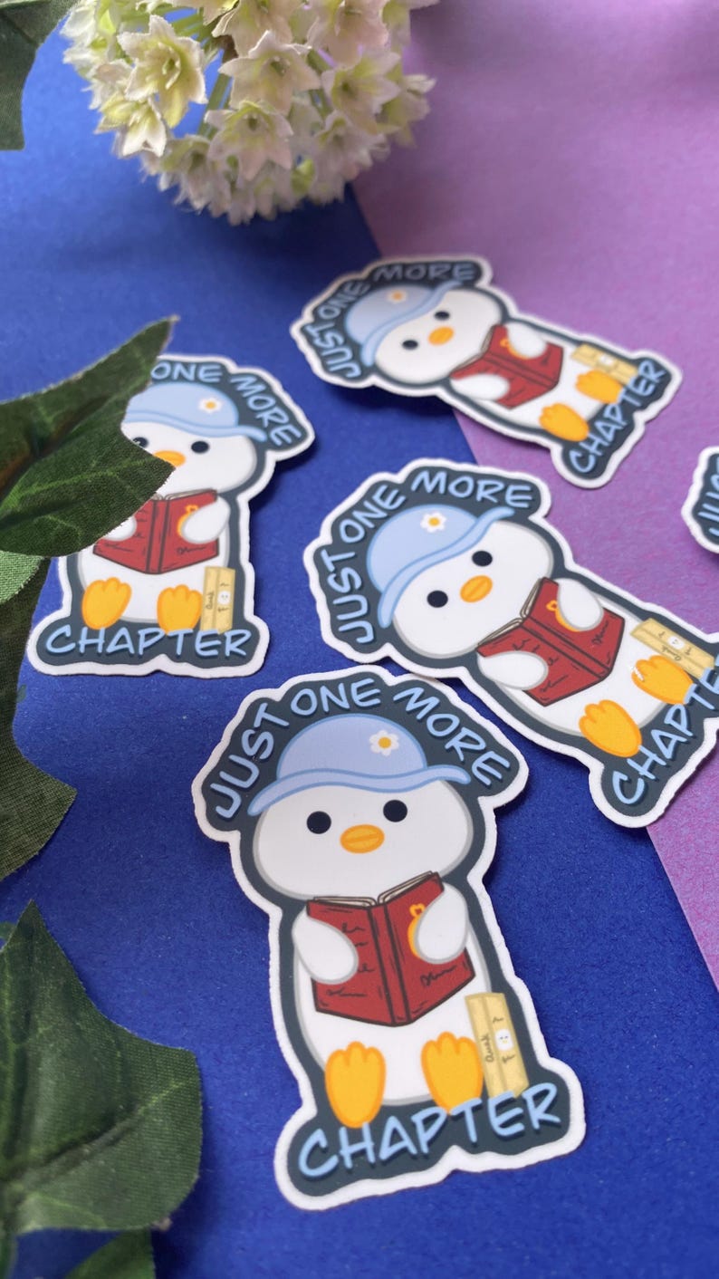 May include: A collection of stickers featuring a cartoon penguin reading a book. The penguin wears a blue hat with the text "Just One More" and the word "Chapter" is also visible. The stickers are on a blue and purple background.