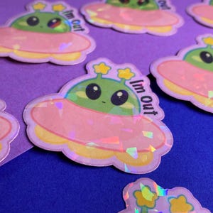 Könnte beinhalten: Eine Sammlung von Aufklebern mit einem Cartoon-Alien in einem UFO. Die Aufkleber haben einen rosa und gelben Rand, einen grünen Alien und den Text "Im Out". Die Aufkleber haben einen holografischen Effekt.