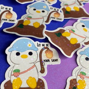 Könnte beinhalten: Eine Sammlung von Aufklebern mit einem Cartoon-Pinguin-Charakter. Der Pinguin trägt einen blauen Hut und hält eine Laterne mit dem Text "Let Me Be Your Light". Die Aufkleber haben ein holografisches, glitzerndes Finish.