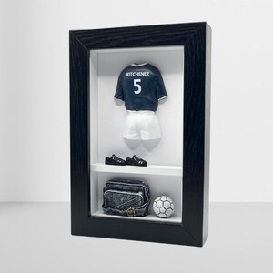 Può includere: Una scatola a ombre incorniciata in nero con sfondo bianco. All'interno della scatola c'è una maglia da calcio in miniatura con il nome "KITCHENER" e il numero "5" sul retro. Ci sono anche scarpini da calcio in miniatura, un pallone da calcio e una piccola borsa nera.