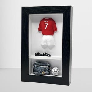 May include: A black framed shadow box featuring a red and white football shirt with the number 7 and the word "BEST" on it. The shirt is displayed with a pair of black football boots, a denim bag, and a white football.