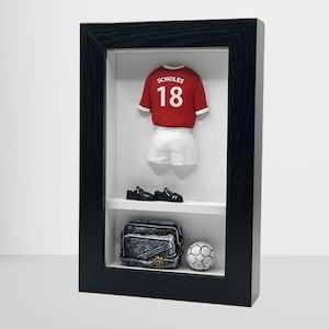 Könnte beinhalten: Eine schwarze gerahmte Schattenbox mit einem rot-weißen Fußballtrikot mit dem Namen "SCHOLES" und der Nummer "18" auf der Rückseite. Das Trikot wird mit einem Fußball, einer schwarzen Tasche und einem Paar schwarzer Schuhe ausgestellt.