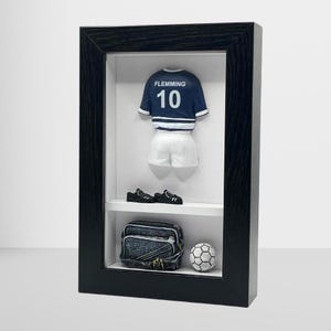 Può includere: Una scatola ombra incorniciata in nero con sfondo bianco. All'interno della scatola c'è una maglia da calcio in miniatura blu e bianca con il nome "FLEMMING" e il numero "10" sul retro. Ci sono anche scarpini da calcio in miniatura neri, una borsa nera e grigia e un pallone da calcio bianco.