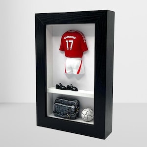 Könnte beinhalten: Eine schwarze gerahmte Schattenbox mit einem rot-weißen Fußballtrikot mit dem Namen "Garnacho" und der Nummer "17" auf der Rückseite. Das Trikot wird mit einem Paar schwarzer Fußballschuhe, einer schwarzen Tasche und einem weißen Fußball präsentiert.