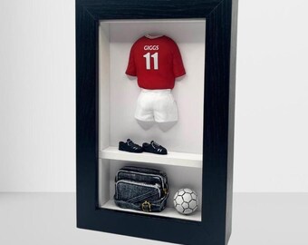 以下が含まれることがあります： 「GIGGS 11」と書かれた赤いサッカーユニフォーム、白いショートパンツ、黒い靴、デニムバッグ、サッカーボールを収めた額入りディスプレイ。 黒いフレームと白い内装がサッカーのテーマを引き立てています。