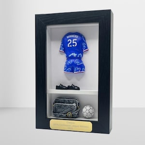 May include: A framed display with a miniature blue football kit, black shoes, a denim bag, and a football. The frame includes a gold plaque that reads "Chelsea UEFA Conference League Winners 2023."
