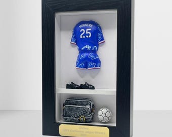 以下が含まれることがあります： 青いサッカーのミニチュアユニフォーム、黒い靴、デニムバッグ、サッカーボールが入った額入りディスプレイ。 フレームには「Chelsea UEFA Conference League Winners 2023」と書かれた金のプラークが含まれています。