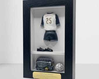 以下が含まれることがあります： 額縁入りのディスプレイ。背番号25のミニチュアサッカーユニフォーム、ショートパンツ、クリート。デニムバッグとサッカーボールも展示。下部の金色の銘板には「Tottenham Hotspur UEFA Europa League Winners 2023」と書かれています。