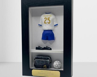 以下が含まれることがあります： 番号25のミニチュアサッカーユニフォーム、青いショーツ、黒いクリートのペアが特徴の額入りディスプレイ。 下には、黒とグレーのバッグとサッカーボールがあります。 金色のプラークには「The Lionesses Champions of Europe 2025」と書かれています。