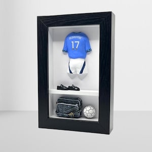 Può includere: Una scatola ombra incorniciata in nero con una maglia da calcio blu in miniatura con il nome "De Bruyne" e il numero "17" sul retro. La maglia è esposta con un pallone da calcio in miniatura, una borsa in miniatura e scarpe nere in miniatura.