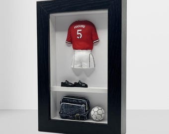 以下が含まれることがあります： 「MAGUIRE 5」と書かれた赤いサッカーのユニフォーム、白いショートパンツ、黒い靴、デニムバッグ、サッカーボールが黒いフレームに収められたディスプレイ。アイテムは棚に配置されています。