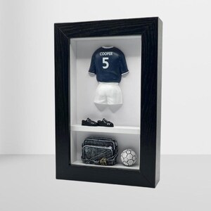 Può includere: Una scatola ombra incorniciata in nero con una miniatura di divisa da calcio. La maglia blu navy ha "COOPER" e il numero "5". L'esposizione include pantaloncini bianchi, scarpe da calcio nere, una borsa da viaggio e un pallone da calcio.