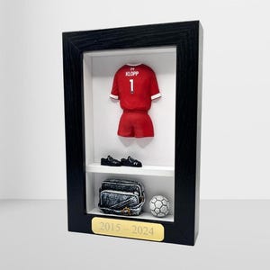 Op de afbeelding: Een zwarte ingelijste schaduwdoos met een rood voetbaltenue met de naam "KLOPP" en het nummer "1" op de rug. Het tenue wordt getoond met een paar zwarte voetbalschoenen, een zwarte voetbaltas en een witte voetbal. De onderkant van de schaduwdoos heeft een gouden plaquette met de tekst "2015 - 2024".
