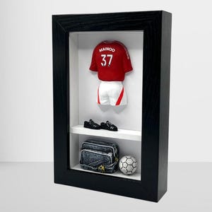 Könnte beinhalten: Eine schwarze gerahmte Schattenbox mit einem rot-weißen Fußballtrikot mit dem Namen "Mainoo" und der Nummer "37" auf der Rückseite. Das Trikot wird mit einem Paar schwarzer Fußballschuhe, einer schwarzen und blauen Tasche und einem weißen Fußball präsentiert.