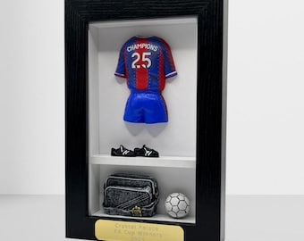 以下が含まれることがあります： 番号25のミニチュアサッカーユニフォーム、青いショーツ、黒いクリートを備えた額入りのディスプレイ。サッカーボールとグレーのバッグも表示されています。下部の金色のプラークには「Crystal Palace FA Cup Winners 2025」と書かれています。