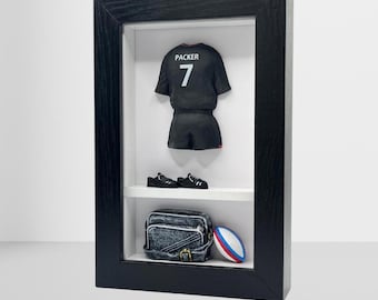 以下が含まれることがあります： 「PACKER」と番号「7」がプリントされた黒いラグビージャージを額装したディスプレイ。ミニチュアの黒い靴、デニムバッグ、赤、白、青のストライプが入ったラグビーボールも含まれています。