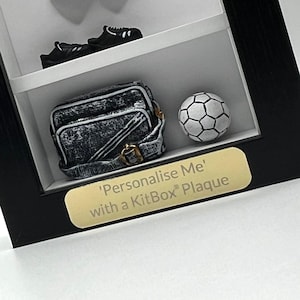 Puede incluir: Una caja de sombra negra con un balón de fútbol, una pequeña bolsa y dos pares de zapatos. La caja tiene una placa dorada que dice "'Personalízame' con una placa KitBox"