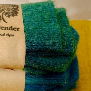Könnte beinhalten: Ein Stapel türkisfarbener und blauer Garnknäuel mit einem braunen Papieretikett mit der Aufschrift "ender" und "cal dyer". Das Garn liegt auf einer gelben Oberfläche.
