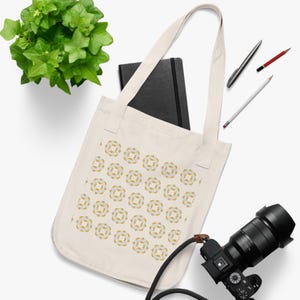 Puede incluir: Bolso tote color crema con un estampado floral repetido en amarillo y blanco. El bolso tiene asas largas y contiene un cuaderno negro. También se ven una cámara, bolígrafos y una planta.