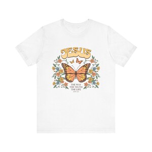 Puede incluir: Camiseta blanca con un diseño de mariposa y la palabra "JESUS" en letras amarillas. La mariposa es naranja y negra, rodeada de flores. El texto "THE WAY THE TRUTH THE LIFE" está debajo de la mariposa.