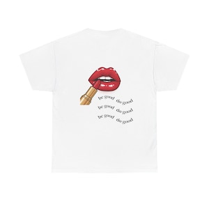 Puede incluir: Camiseta blanca con un gráfico de labios rojos y un lápiz labial dorado. Las palabras "be good do good" están impresas debajo en un diseño curvo. Una camiseta casual y elegante.