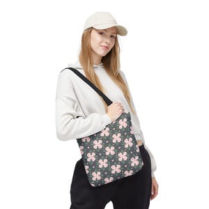 Puede incluir: Un bolso tote con un estampado floral. El bolso presenta un fondo gris oscuro con un patrón repetido de flores rosas, pequeñas hojas verdes y pequeños corazones rosas. Tiene una correa negra para el hombro.
