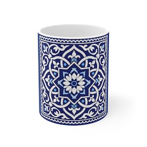 Puede incluir: Una taza de cerámica blanca con un patrón geométrico azul y blanco. El diseño presenta un motivo central en forma de estrella con elementos florales, enmarcado por un borde de patrones arremolinados. La taza tiene un interior blanco.