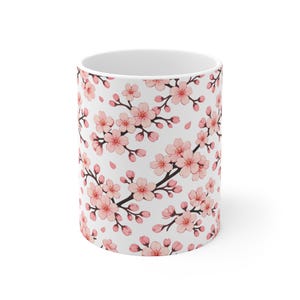 Puede incluir: Taza de cerámica blanca con un patrón repetido de ramas de flores de cerezo rosas y pétalos cayendo. El diseño incluye flores rosas detalladas y ramas marrones oscuras.