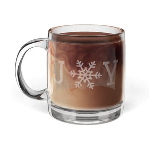 May include: A clear glass mug with a handle, filled with a beverage that has a swirl of cream and a dark brown liquid. The word "JOY" is etched on the front, with a snowflake replacing the "O".