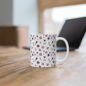 Puede incluir: Taza de cerámica blanca con un estampado floral. El diseño presenta flores moradas, rosas y amarillas con hojas verdes sobre un fondo crema. La taza tiene un asa curva y está sobre una superficie de madera.