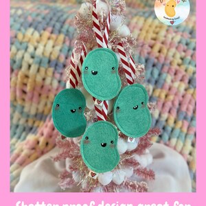 May include: Four teal bean-shaped ornaments with smiling faces hang from a small pink Christmas tree decorated with candy cane striped ribbons. Each ornament has a small heart tag. The ornaments are shatterproof.