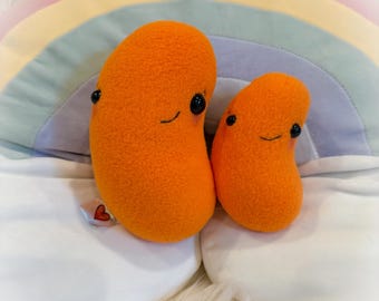 Puede incluir: Dos peluches naranjas con forma de judía, con ojos negros y sonrisas cosidas. Descansan sobre una almohada blanca con forma de nube frente a un arco iris. Los peluches están hechos de un material suave y texturizado.