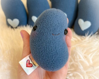 以下が含まれることがあります： 黒いビーズの目と縫い付けられた笑顔が付いた、青い豆の形をしたぬいぐるみのおもちゃのコレクション。白いハートのアップリケが付いているものもあります。1つは手に持たれており、「BeanTown Buddies」とハートのデザインが書かれたタグが付いています。